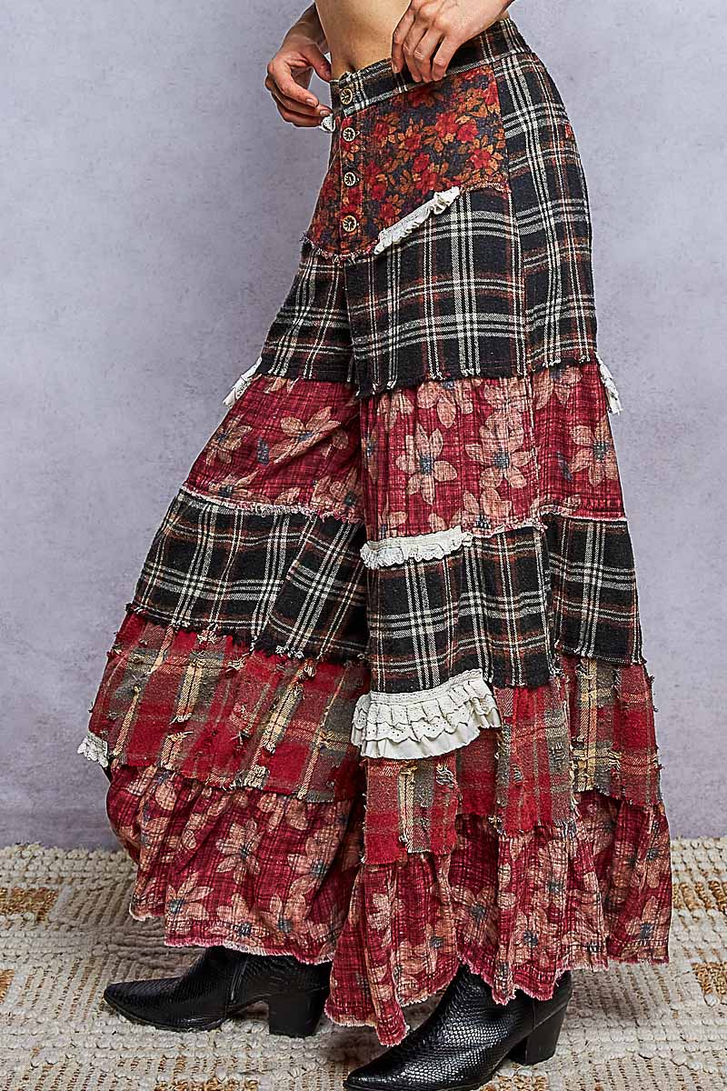 A long, tiered patchwork skirt with red floral and black plaid patterns, featuring white lace ruffles and a buttoned waist, modeled with black boots against a textured gray background.