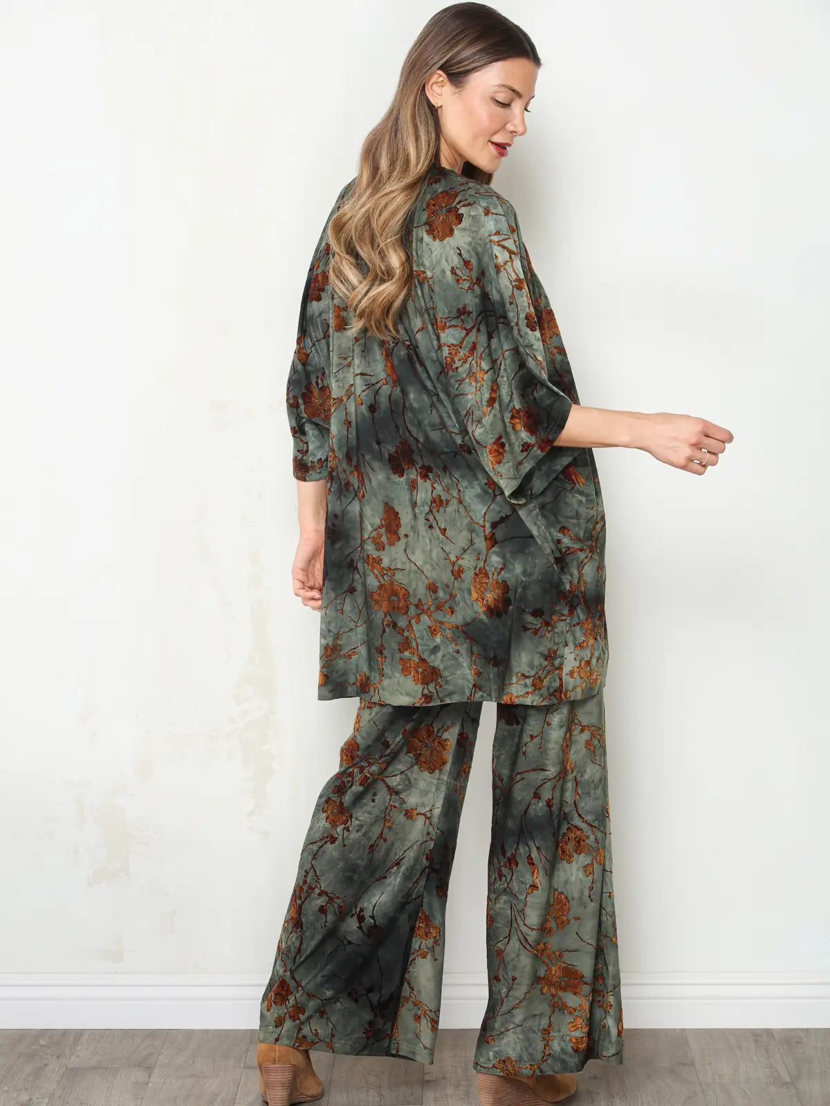 A woman modeling a matching two-piece lounge set with a dark green and brown floral branch print, shown from a side-back angle against a white wall.