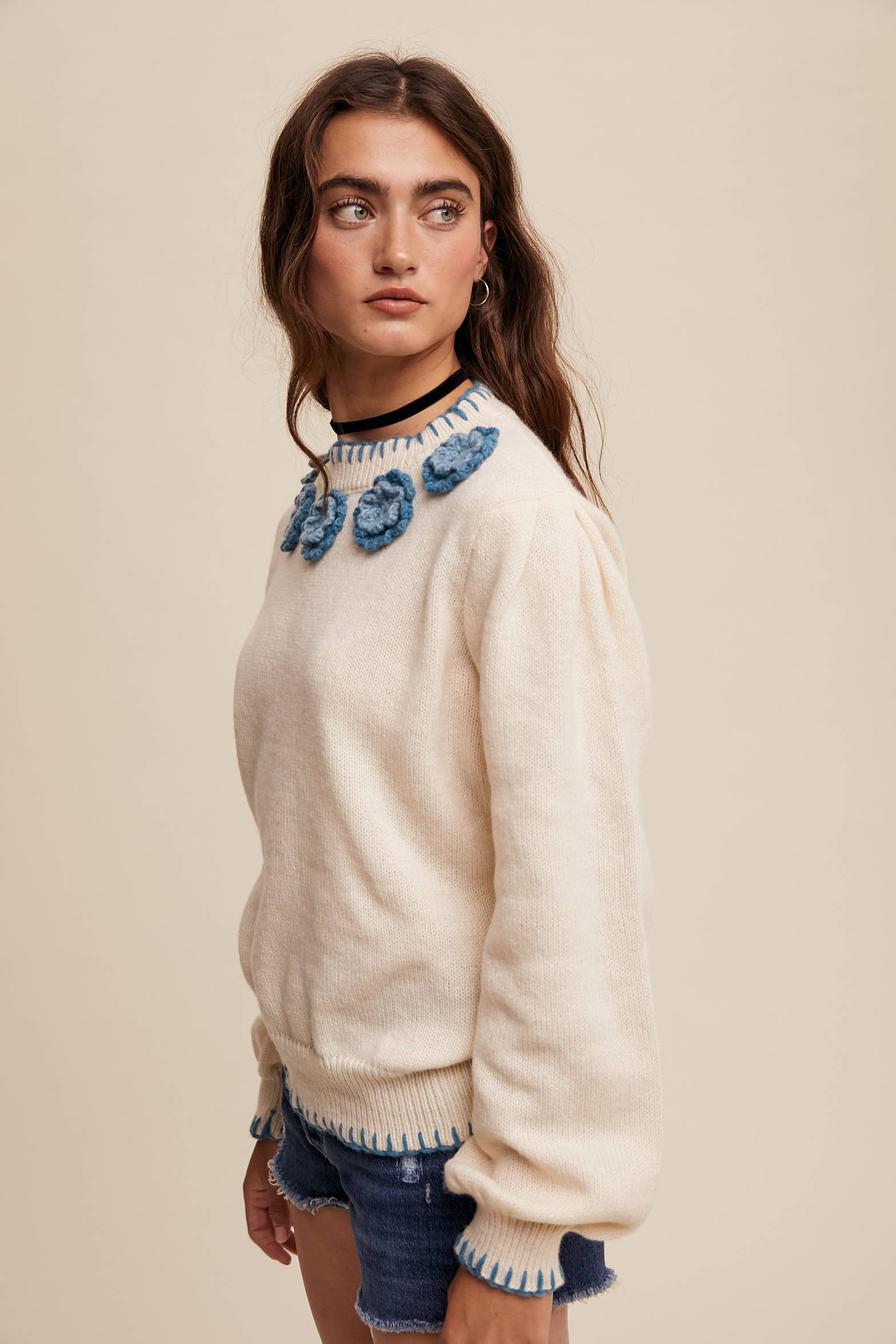 Side view- Cream sweater with blue crochet flower appliqués at neckline, blue whipstitch trim at cuffs and hem, long sleeves; styled with frayed denim shorts.
