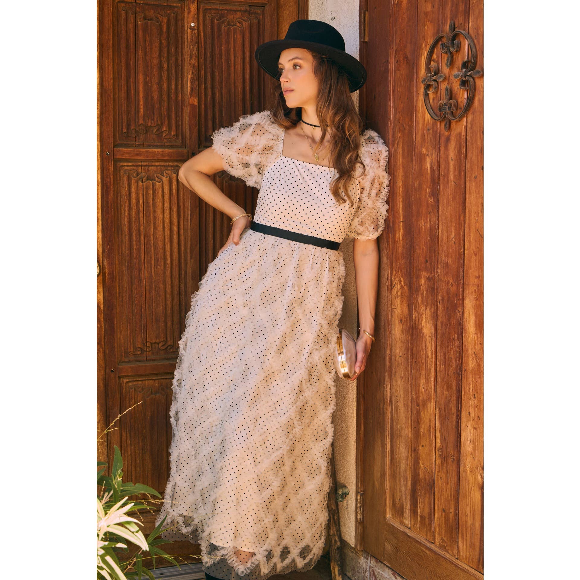 Front view — cream polka-dot tulle midi dress with puff sleeves and black belt; styled with black hat, ankle boots, pearl clutch; leaning by wooden doors.