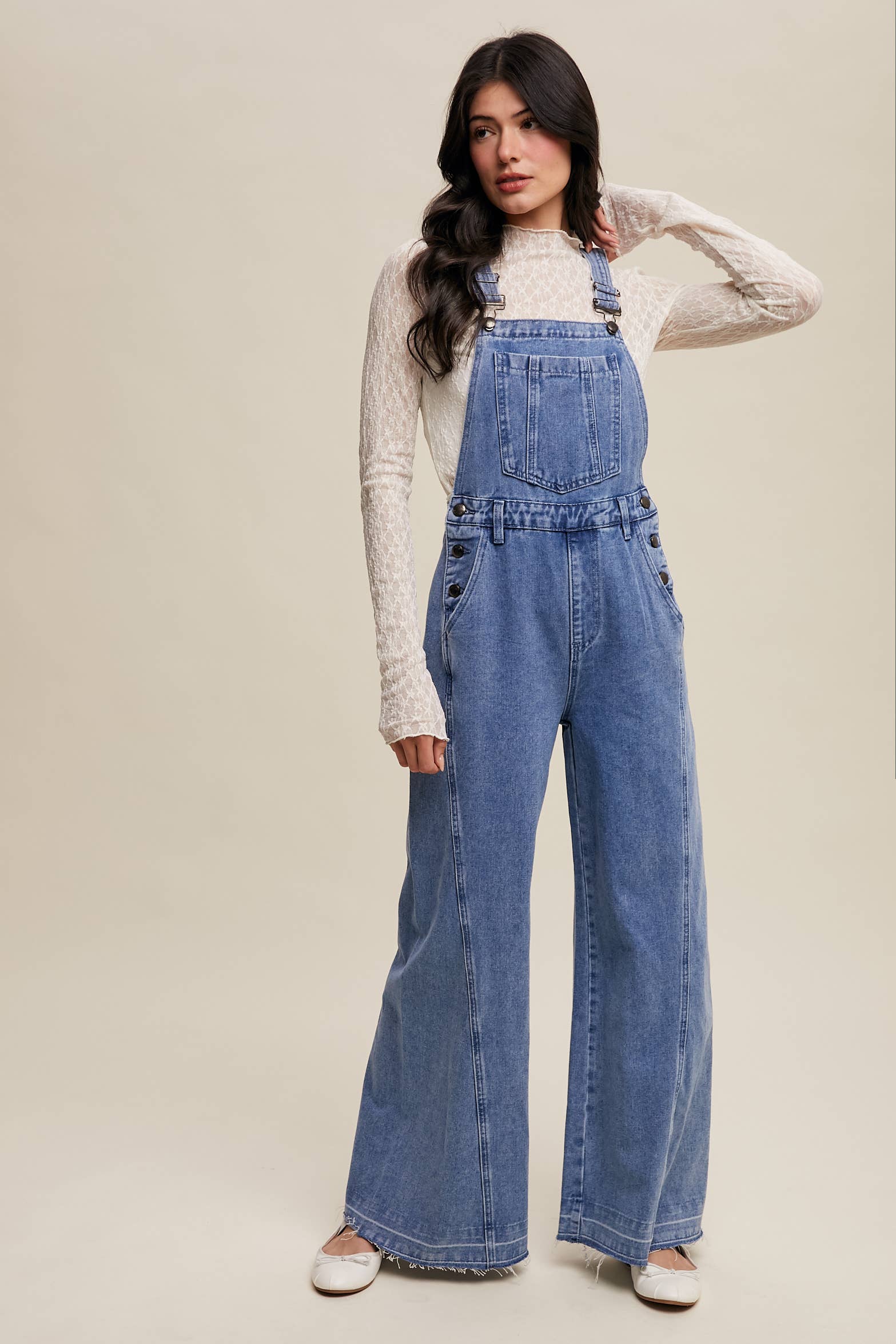 Light-wash wide-leg denim overalls with adjustable straps, front bib pocket, side buttons, and slant pockets, finished with a raw hem and layered over a cream long-sleeve top.