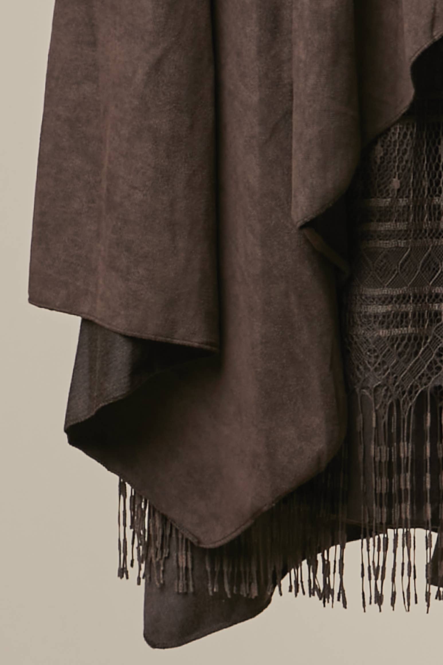 A close-up of a dark brown draped fabric with long fringe, layered over a darker patterned textile, against a plain light beige background.