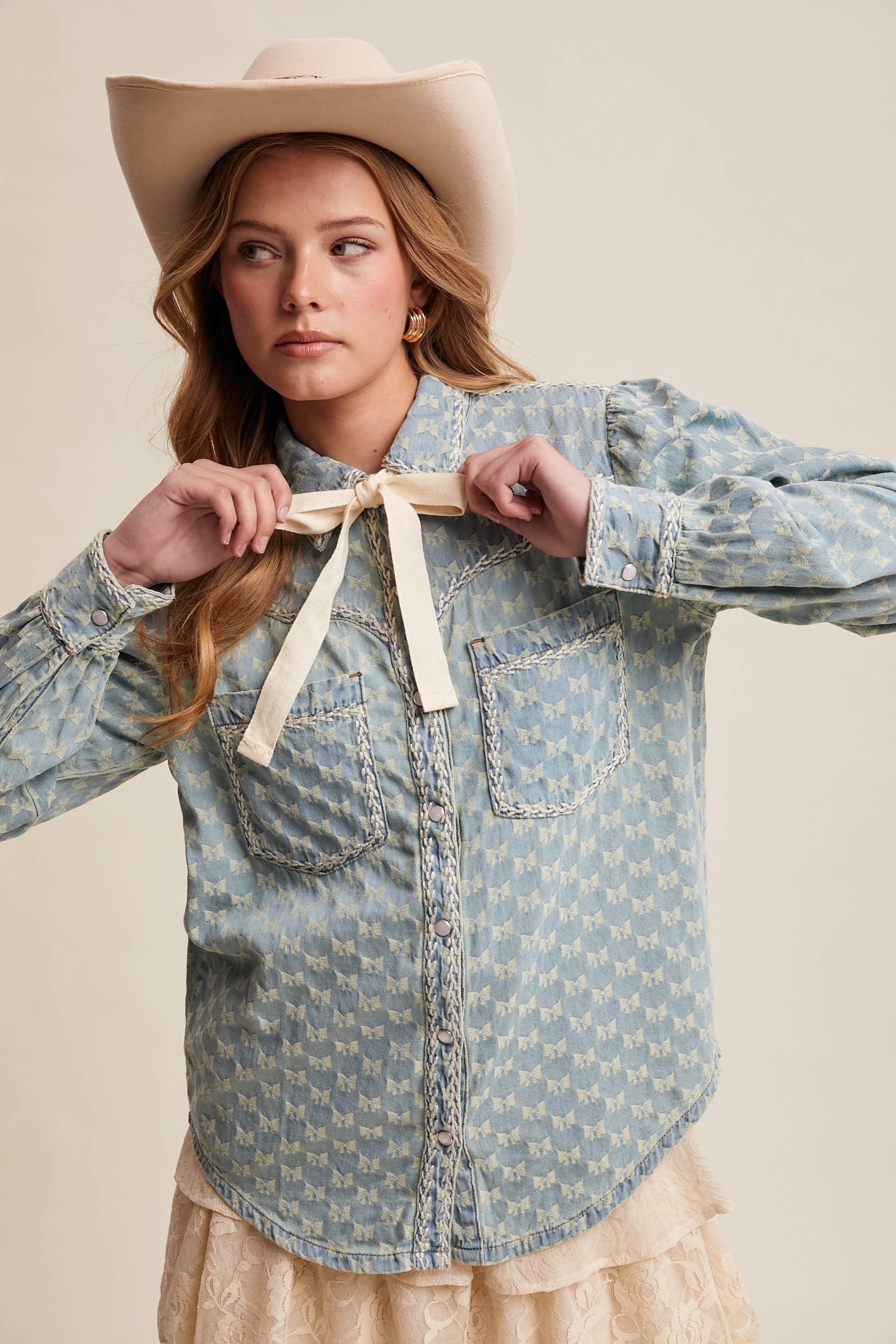 Light-wash denim blouse with cream bow-tie collar, tonal mini-bow pattern, braided contrast stitching, snap front, and two chest pockets.