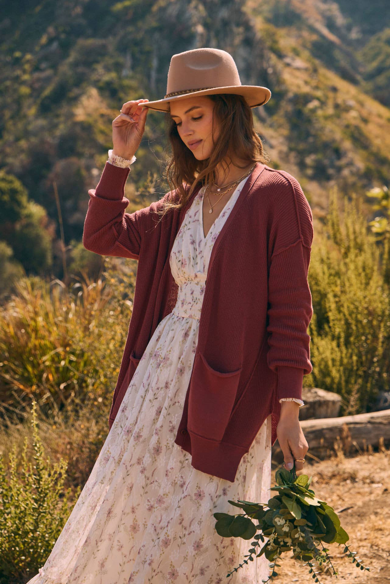 Close-up — mauve knit cardigan layered over cream floral dress with V-neck; tan felt hat; model holding a greenery bouquet outdoors.