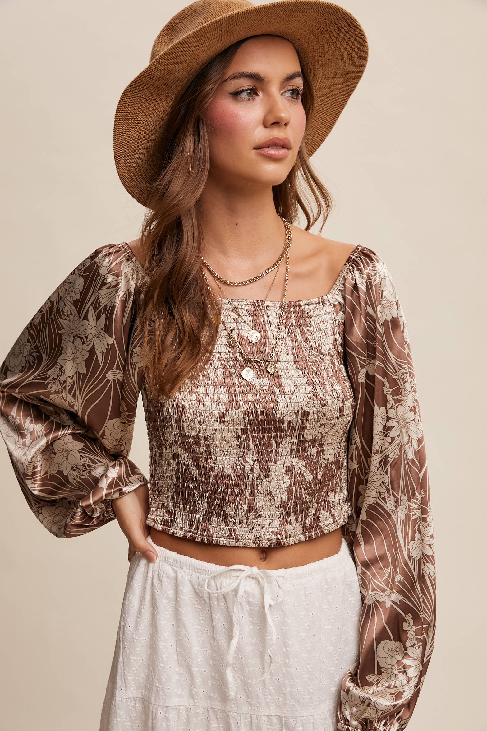 Brown and cream floral smocked blouse with square neckline, puffed long sleeves, and elastic cuffs, styled with layered necklaces and a wide-brim hat.