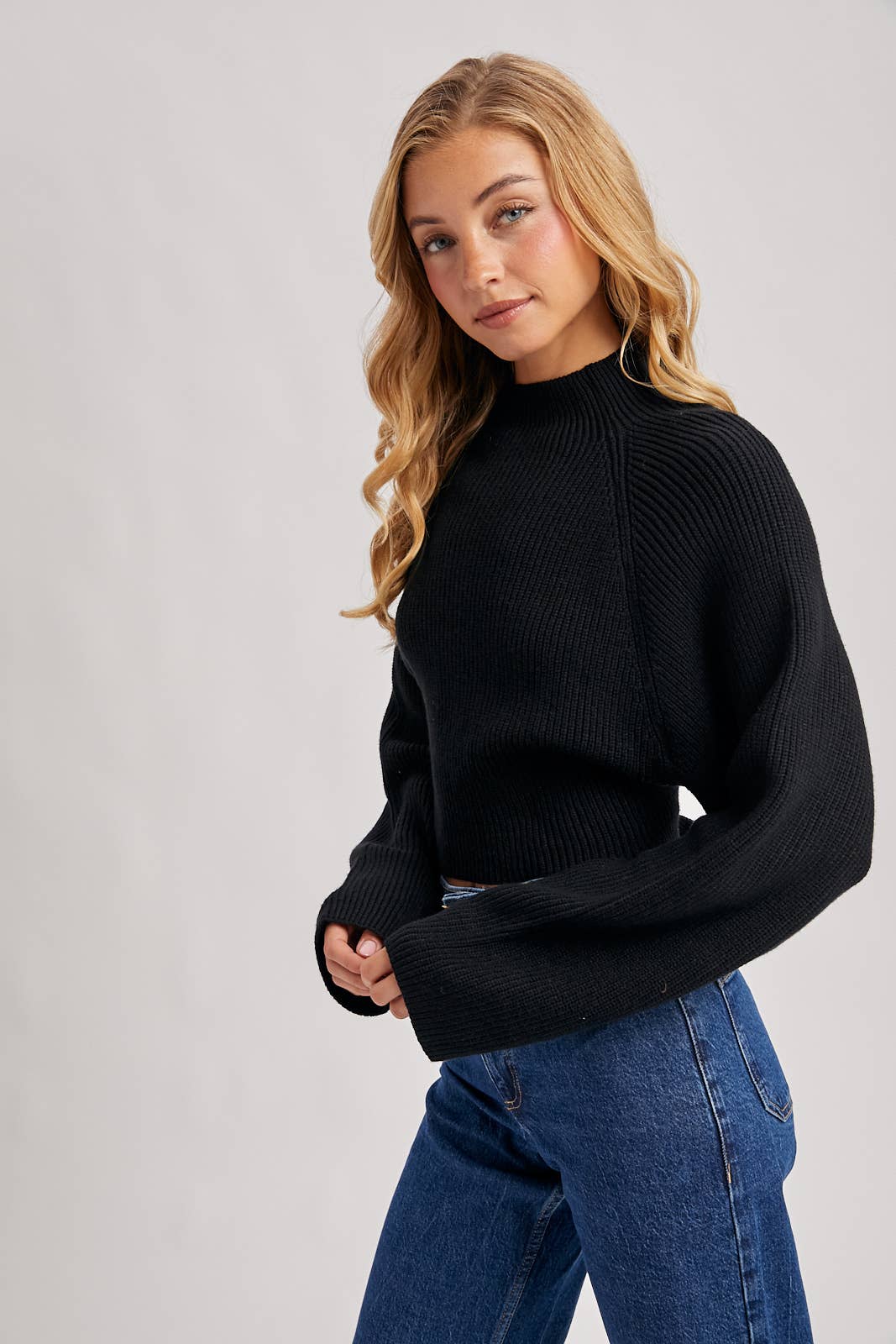 Black ribbed mock-neck sweater with long wide sleeves and a cropped fit, styled with high-rise blue jeans.