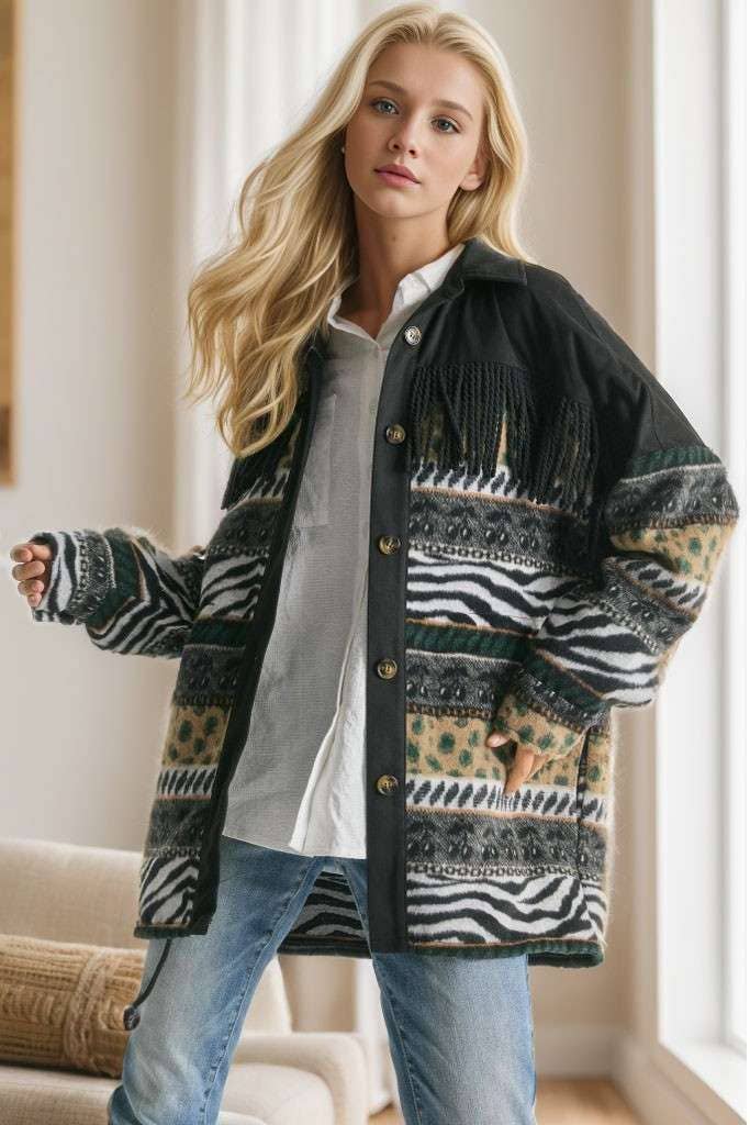 Brown Aztec print shacket with fringe detail, button closure, and patterned sleeves, styled with cream pleated pants while seated on a sofa.