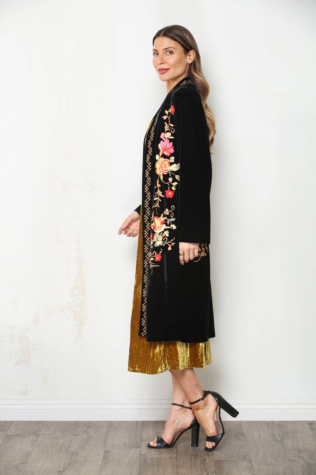Side profile of black velvet floral embroidered duster layered over a gold dress with black block-heel sandals.