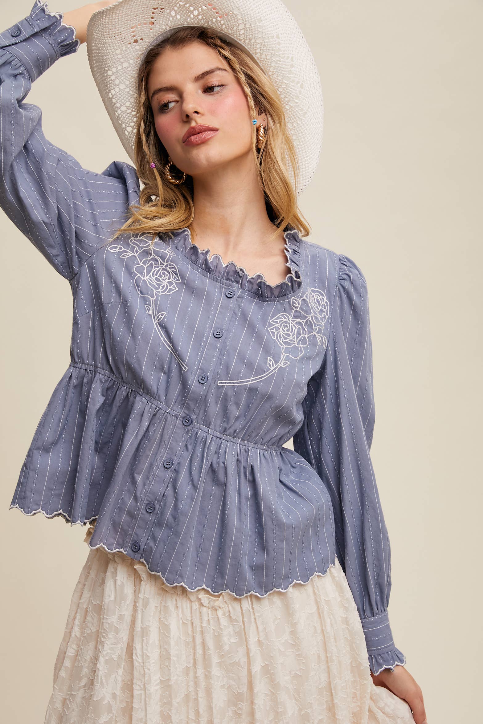 Dusty blue pinstripe blouse with ruffled neckline, white rose embroidery, puff long sleeves, button front, and scalloped peplum hem—styled with a white lace skirt and cowboy hat.