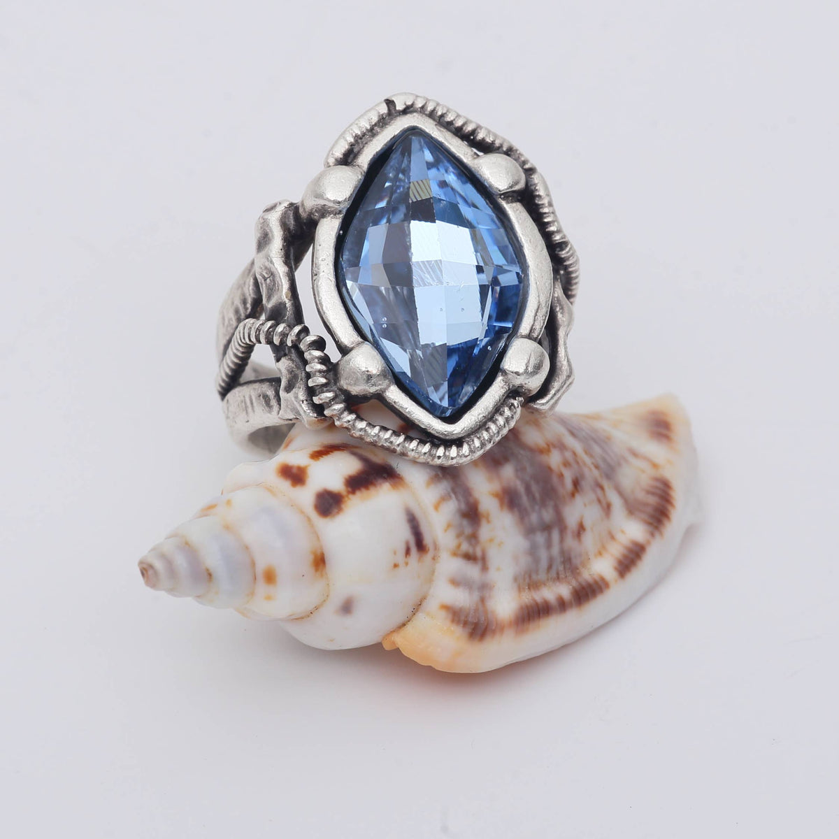 A silver-toned ring with a marquise-cut blue gemstone, displayed on top of a white and brown seashell against a plain white background.