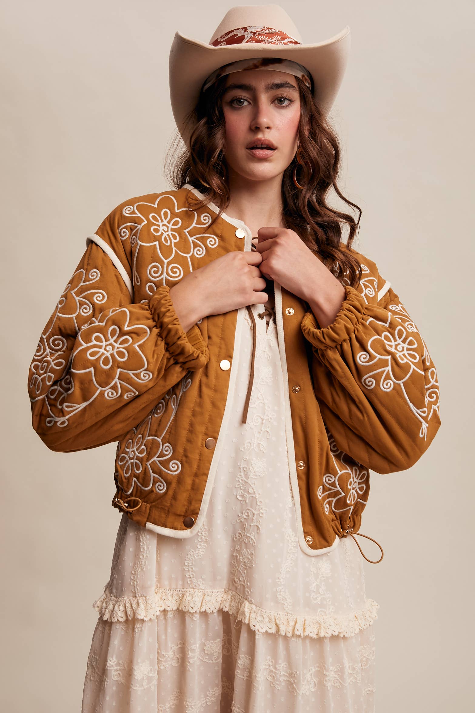 Boho caramel-brown jacket with cream floral swirl embroidery, ivory piping, and snap buttons, styled over a lace tie-front top with a scarf and felt hat outdoors.