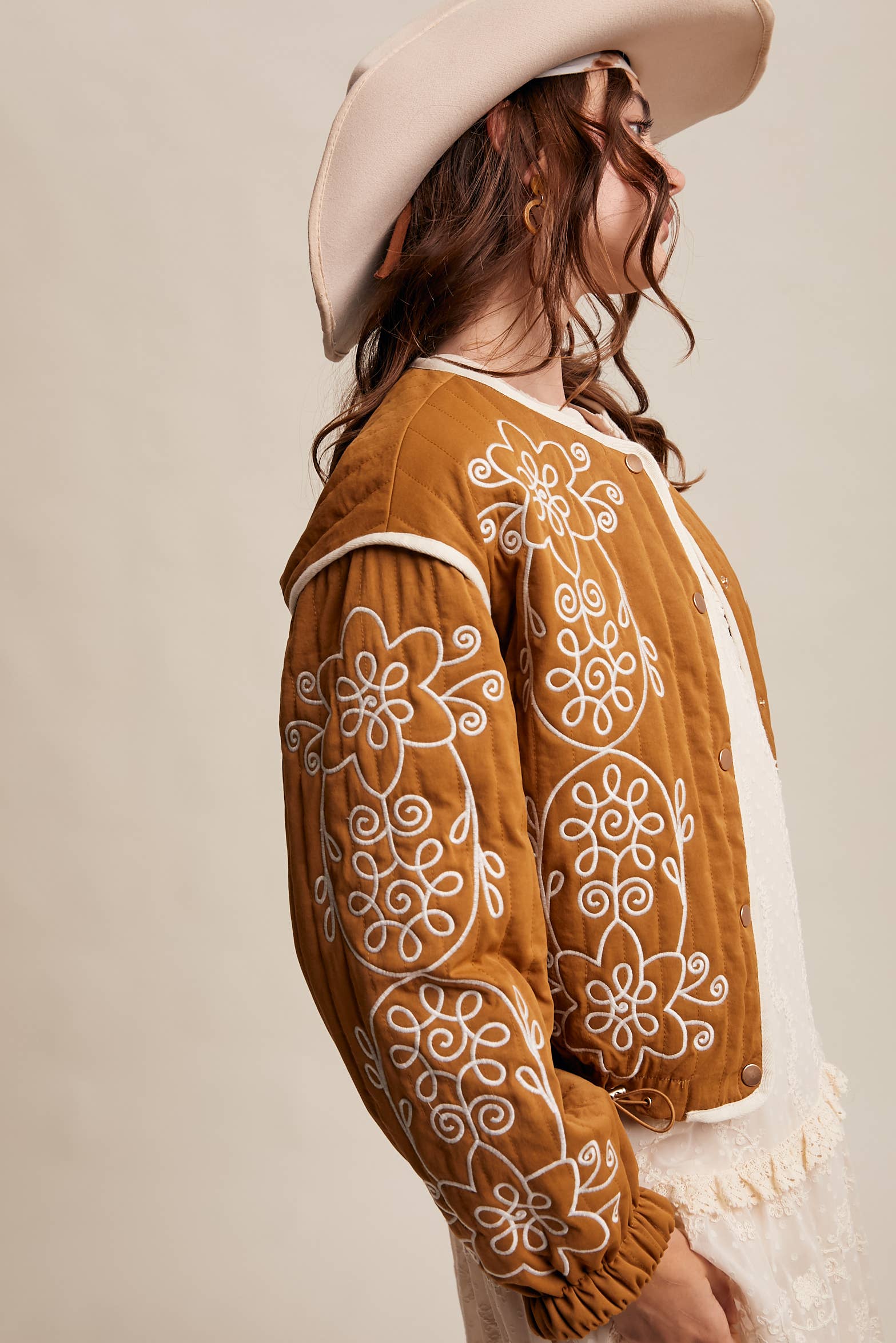 Boho caramel-brown jacket with cream floral swirl embroidery, ivory piping, and snap buttons, styled over a lace tie-front top with a scarf and felt hat outdoors.