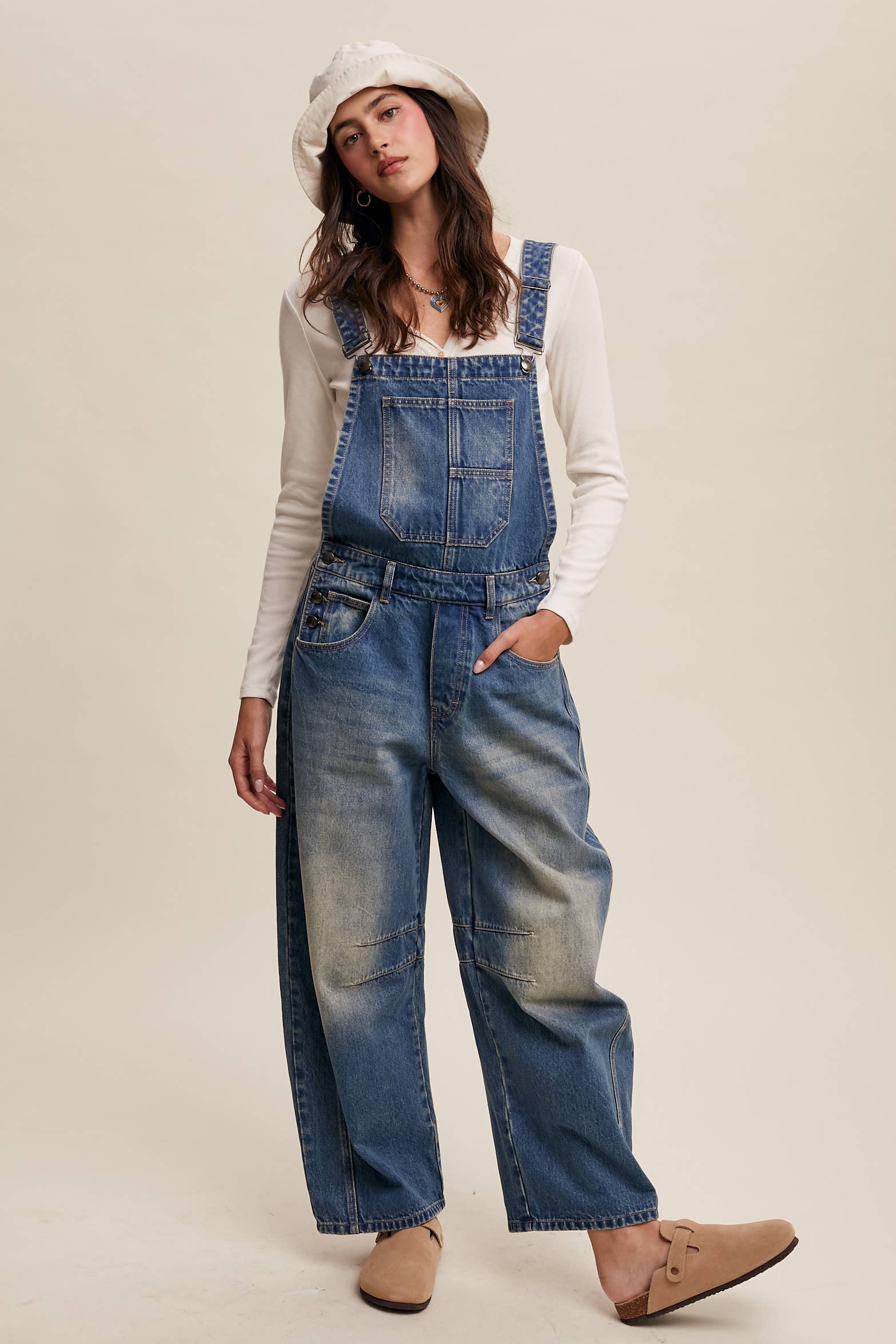 Vintage-inspired blue denim overalls with relaxed wide-leg fit, front chest pocket, and adjustable straps, styled with a white long-sleeve top and neutral clogs.