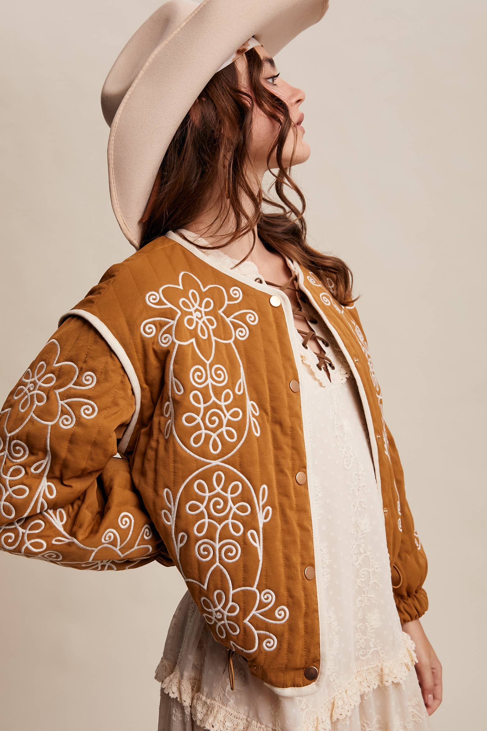 Boho caramel-brown jacket with cream floral swirl embroidery, ivory piping, and snap buttons, styled over a lace tie-front top with a scarf and felt hat outdoors.
