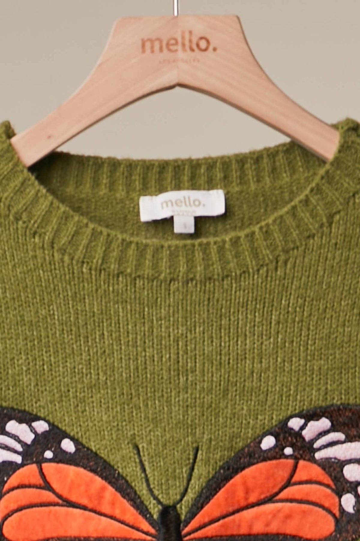 An olive green knit sweater with a large orange and black butterfly applique on the front, hanging on a light wood hanger branded 'mello.'.
