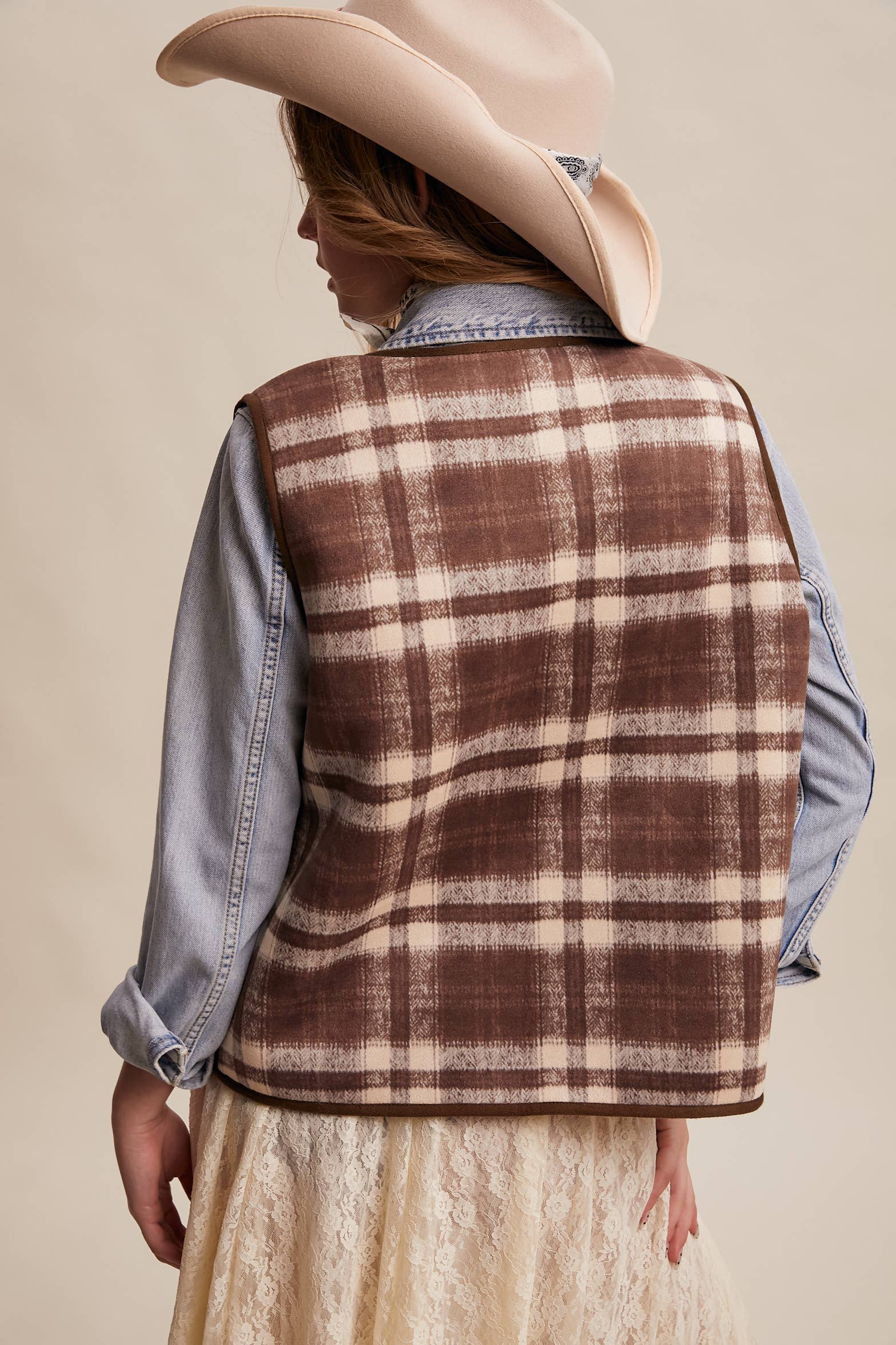 Person wearing a plaid vest over a denim shirt with a beige hat on a beige background