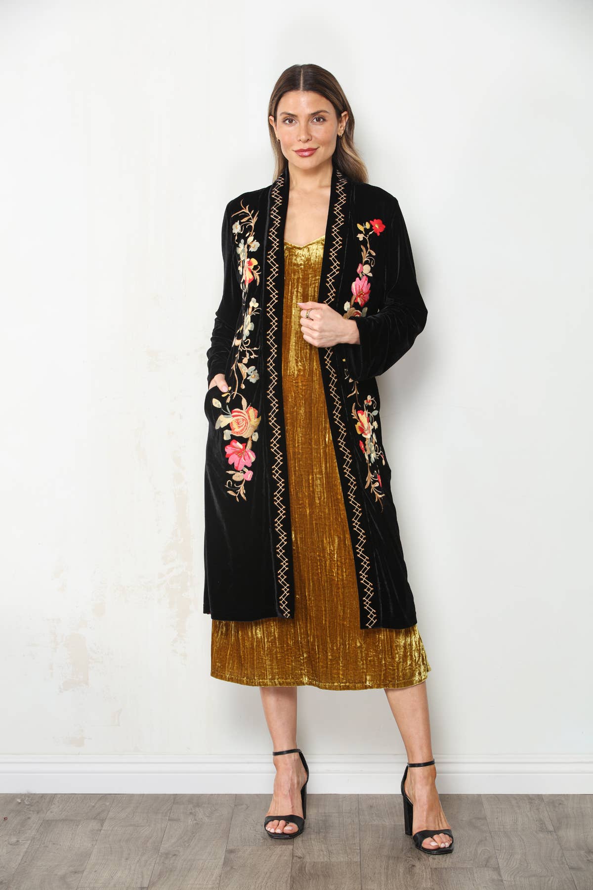 Model wearing black floral embroidered velvet duster with one hand in a pocket, gold dress and black heels.