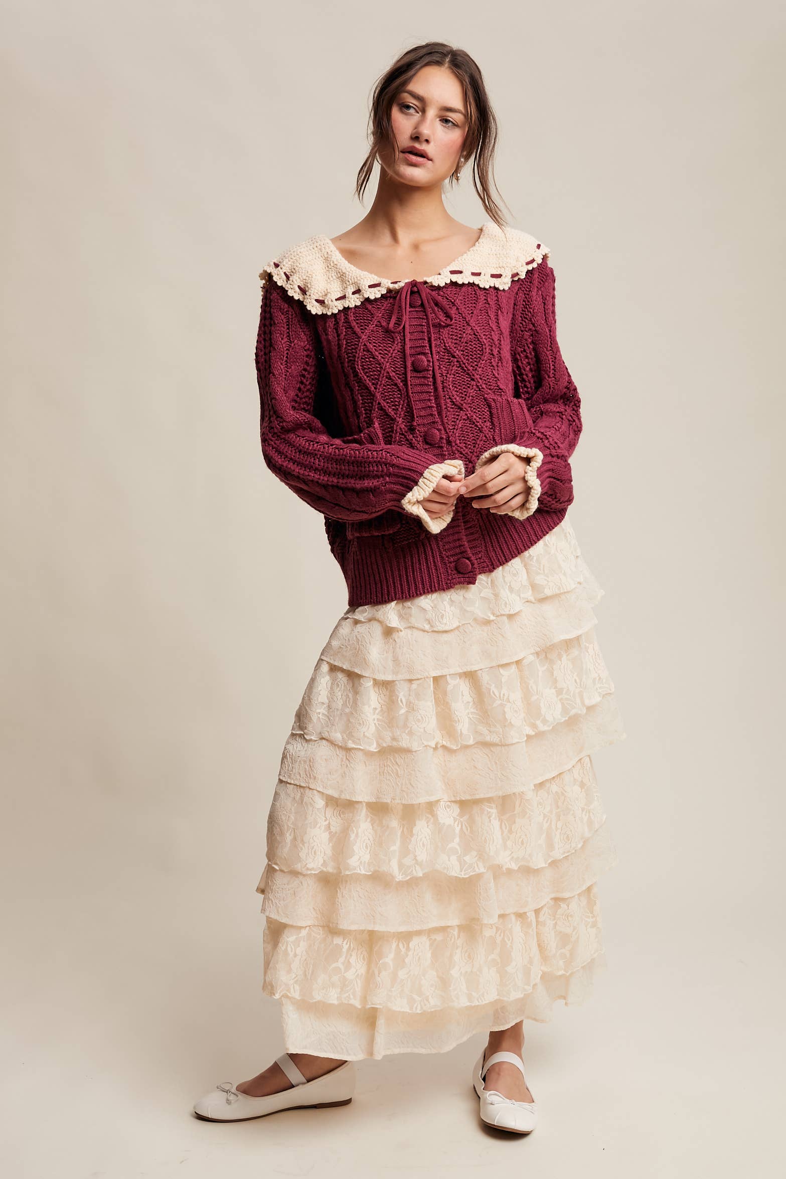 Woman wearing a burgundy cable knit sweater and cream ruffled skirt on a plain background