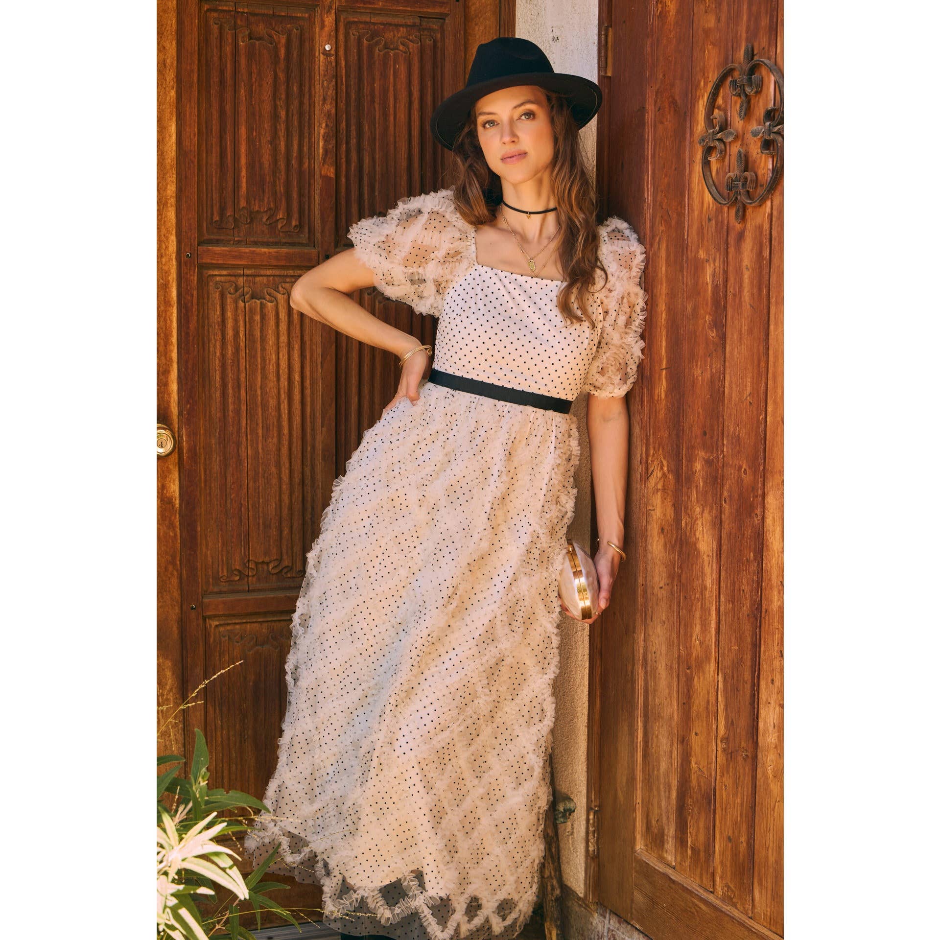 Front view — cream polka-dot tulle midi dress with puff sleeves and black belt; styled with black hat, ankle boots, pearl clutch; leaning by wooden doors.