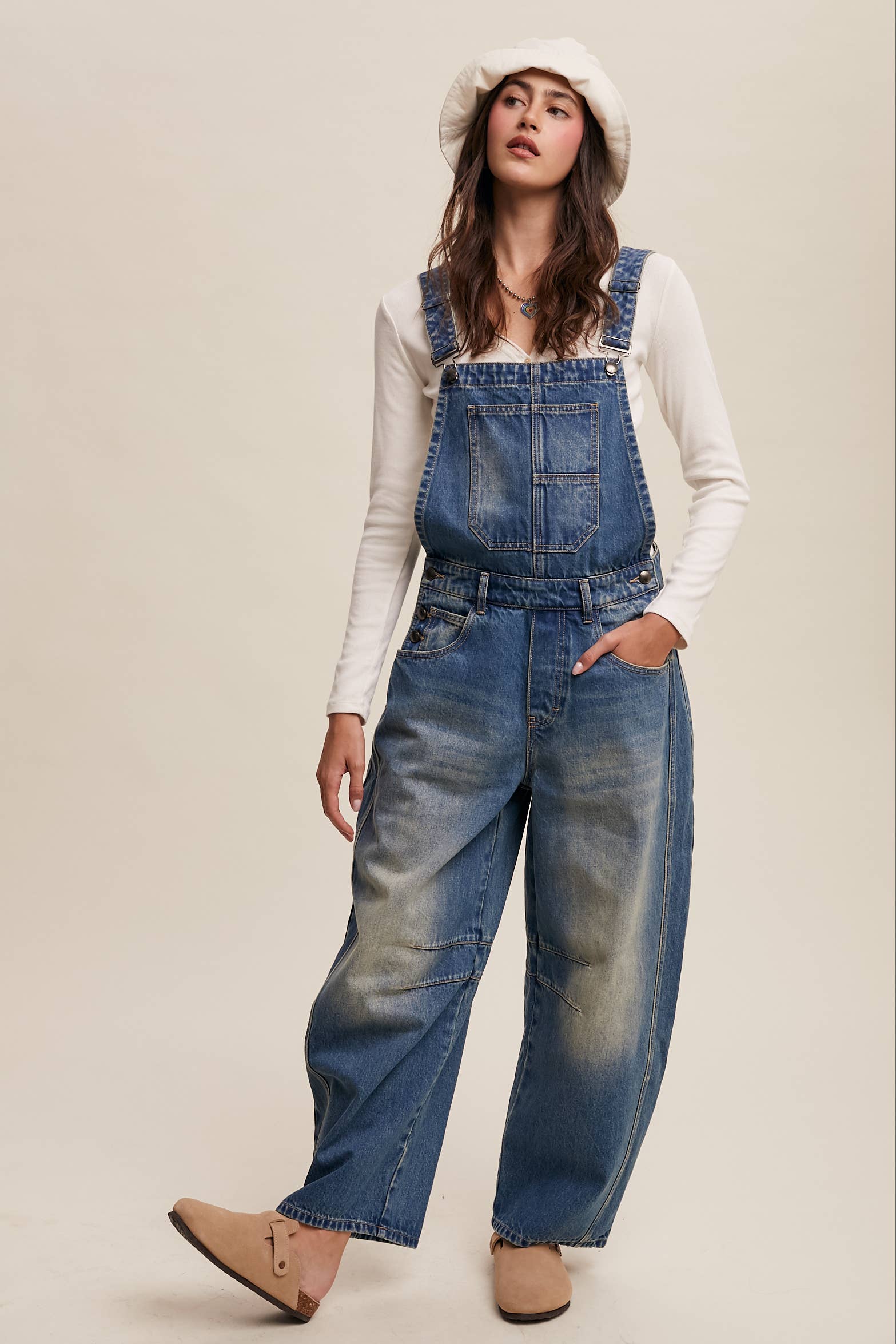 Vintage-inspired blue denim overalls with relaxed wide-leg fit, front chest pocket, and adjustable straps, styled with a white long-sleeve top and neutral clogs.