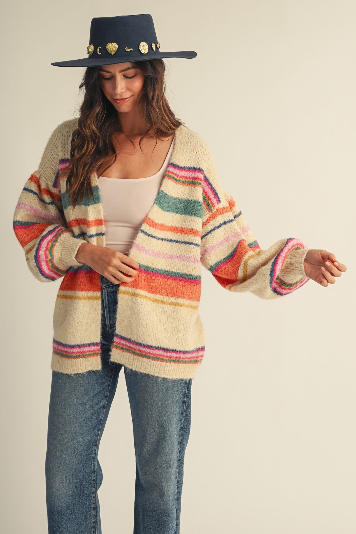 A woman wearing a cream-colored fuzzy knit cardigan with colorful horizontal stripes in pink, orange, green, and blue, paired with a beige tank top, blue jeans, and a dark navy wide-brim hat with gold heart embellishments, set against a plain beige background.