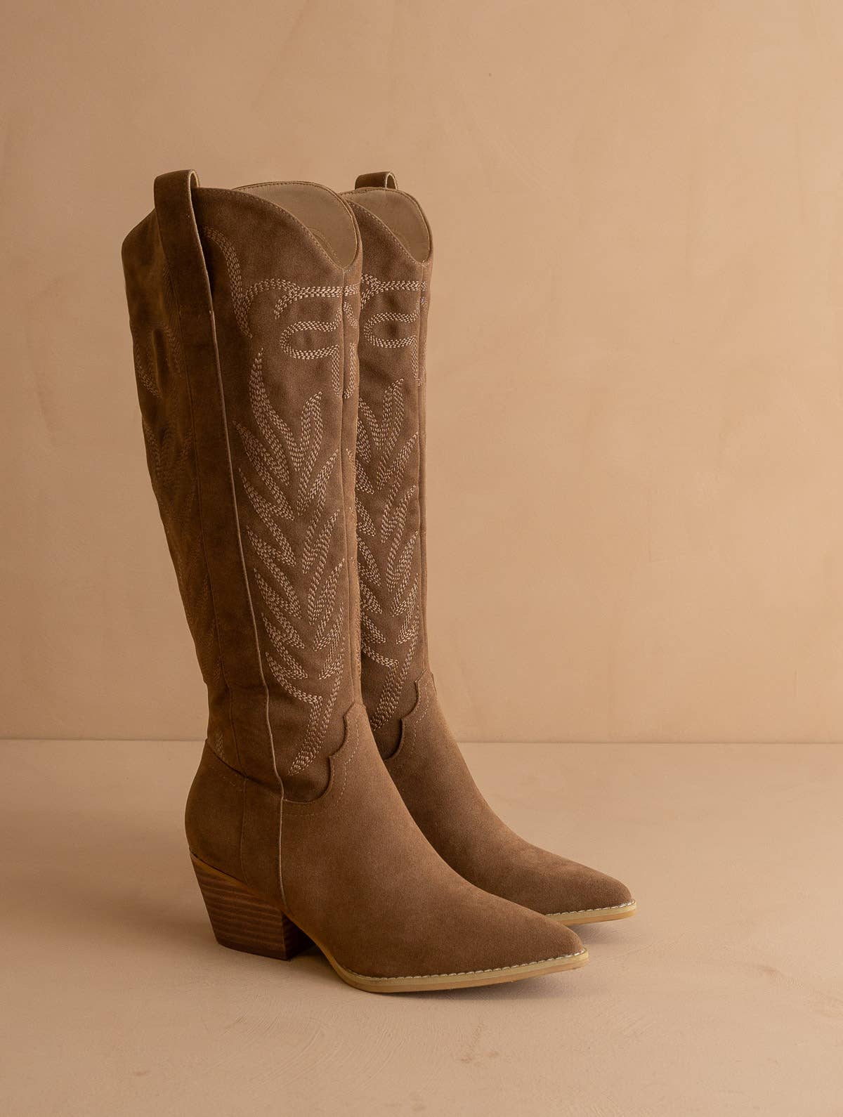 Taupe suede knee-high western boot with stitched shaft, pull tab, pointed toe, and stacked block heel, side view.