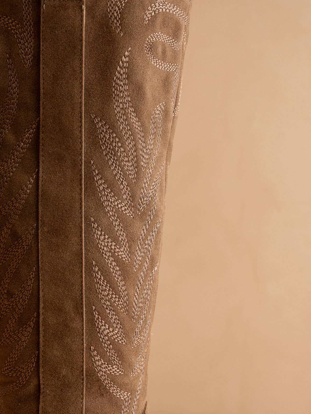 Close-up of taupe suede knee-high western boot shaft with cream decorative stitching detail.