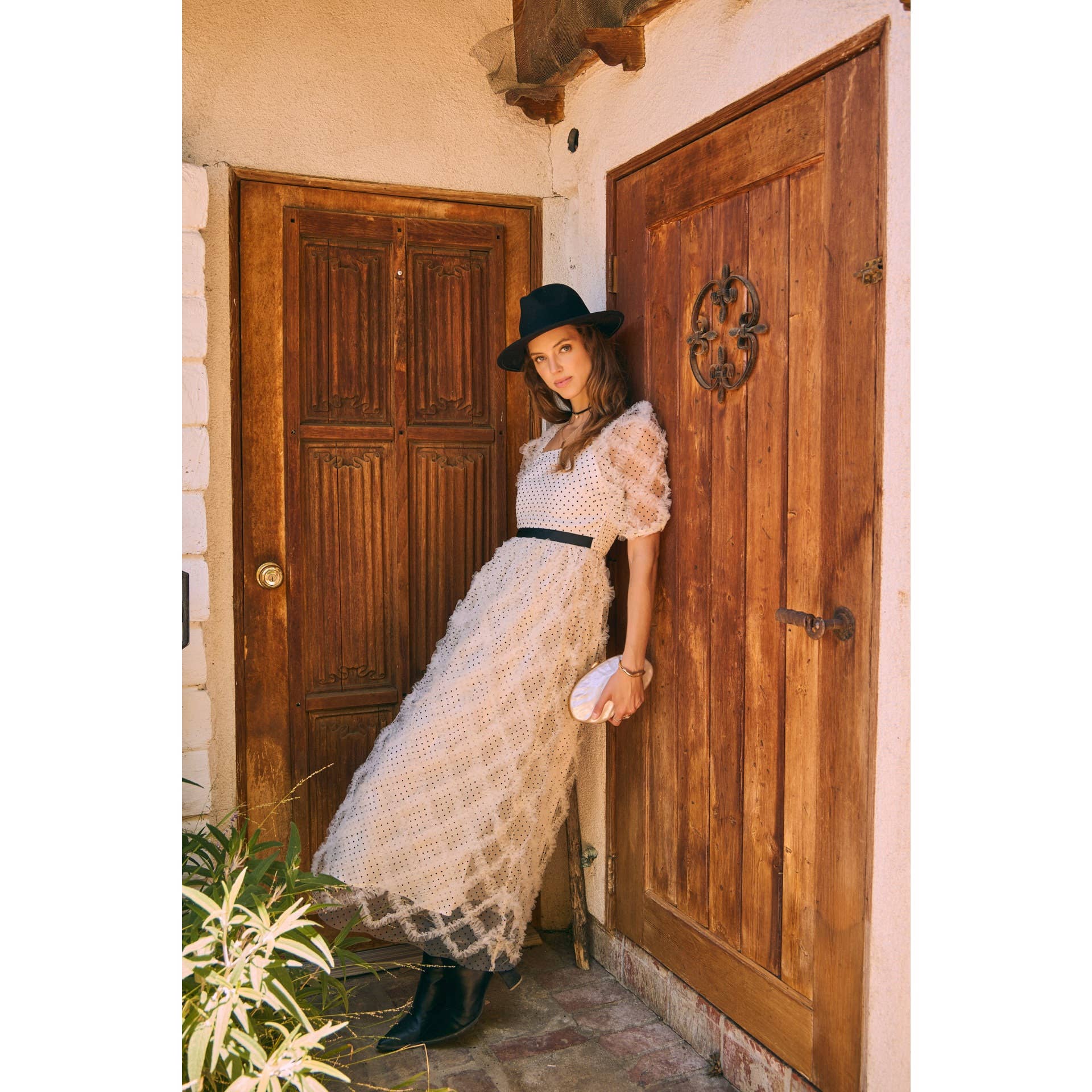Leaning pose — cream polka-dot tulle midi dress with puff sleeves and black belt, styled with black wide-brim hat and ankle boots by a wooden door.