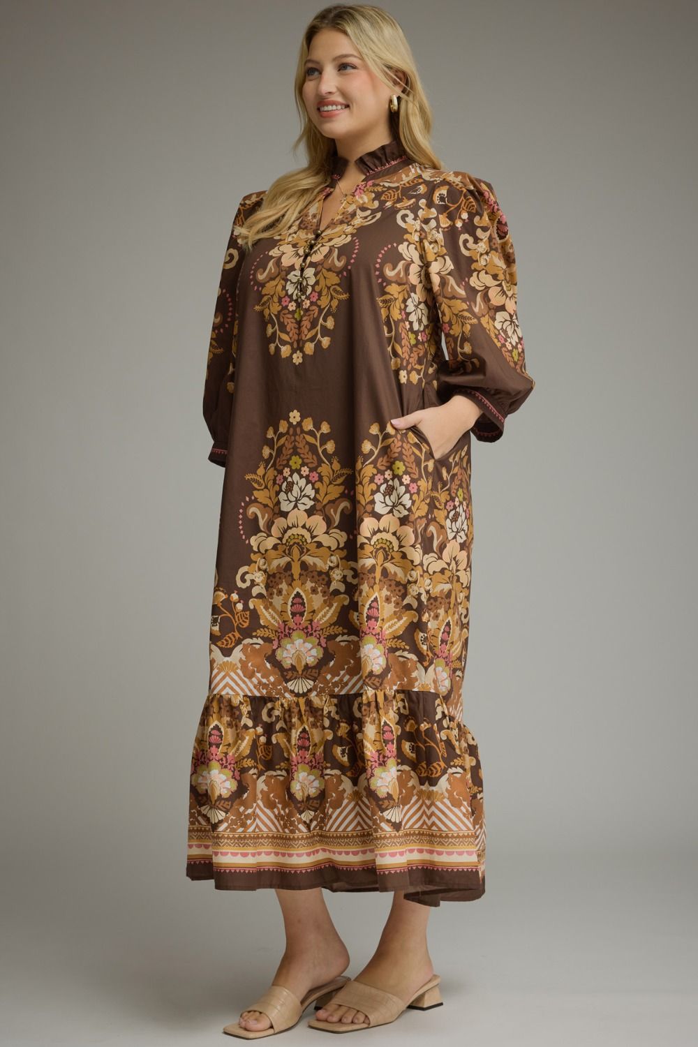 A woman modeling a brown midi dress with an intricate gold, beige, and white floral pattern, standing against a neutral gray studio background.