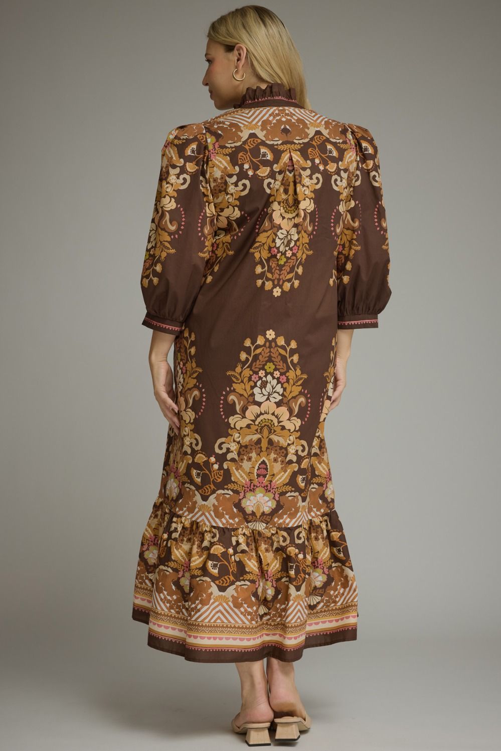 A woman shown from the back wearing a brown midi dress with an ornate beige, gold, and white floral baroque pattern, photographed against a neutral gray studio background.