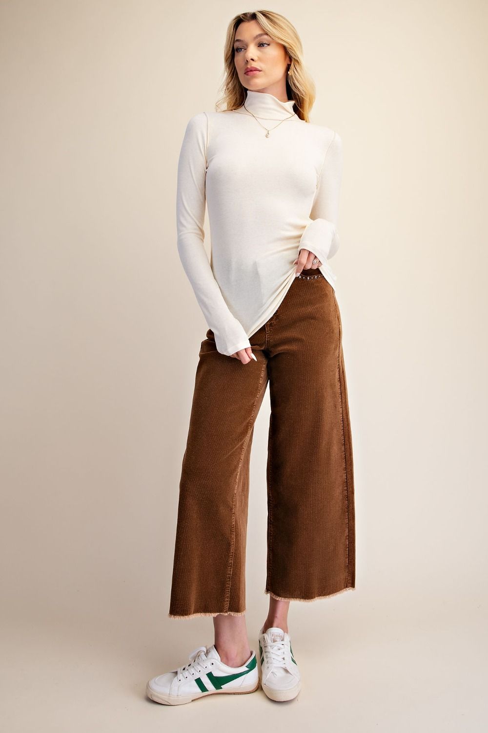 A woman modeling a white long-sleeve mock neck top and brown wide-leg cropped pants, paired with white sneakers, against a solid beige studio background.