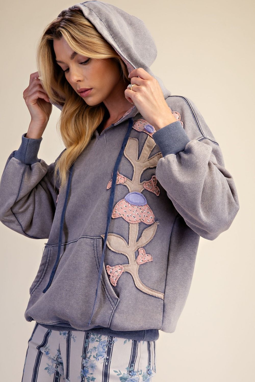 Three-quarter front view of woman wearing slate blue mineral wash hoodie with floral patchwork detail and contrast hood lining, showing kangaroo pocket and dropped shoulder silhouette