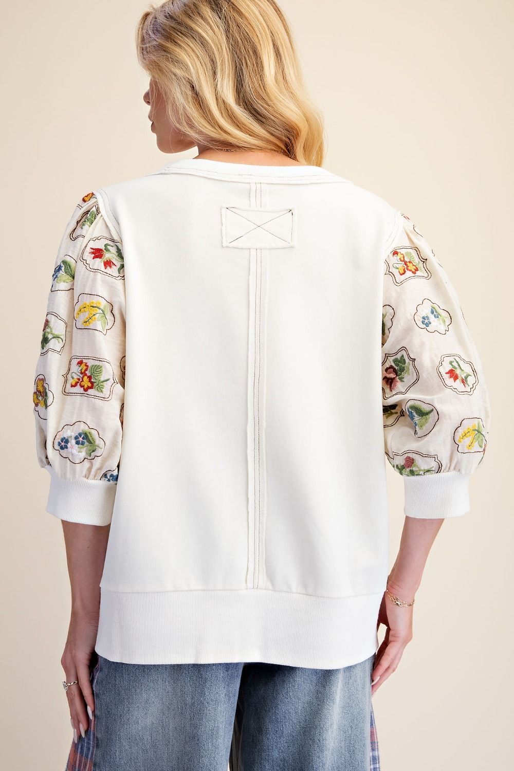 A woman shown from the back wearing a white cropped sweatshirt with colorful embroidered puff sleeves and blue jeans, against a plain beige studio background.