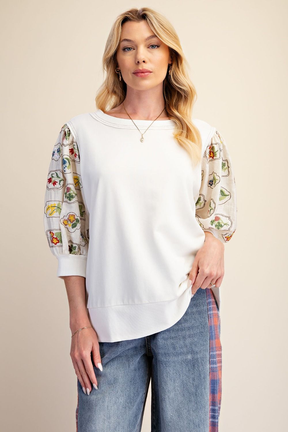 A woman modeling a white crewneck sweatshirt with three-quarter sleeves featuring a colorful floral and butterfly print, paired with blue jeans against a solid beige background.