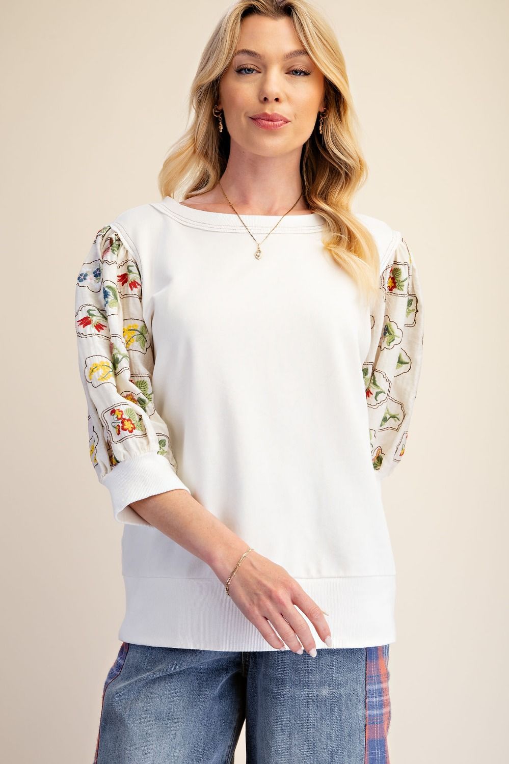 A woman modeling a white crewneck sweatshirt with three-quarter sleeves featuring colorful embroidered floral and vine patterns, paired with blue jeans against a solid beige background.