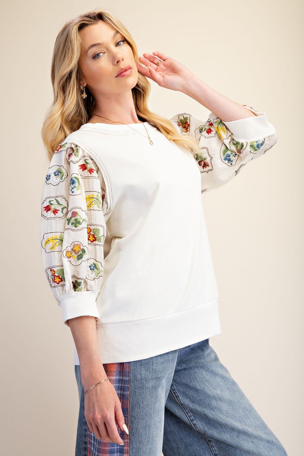 A woman modeling a white crewneck sweatshirt with three-quarter sleeves featuring colorful embroidered fruit and vegetable patterns, paired with blue jeans against a solid beige background.