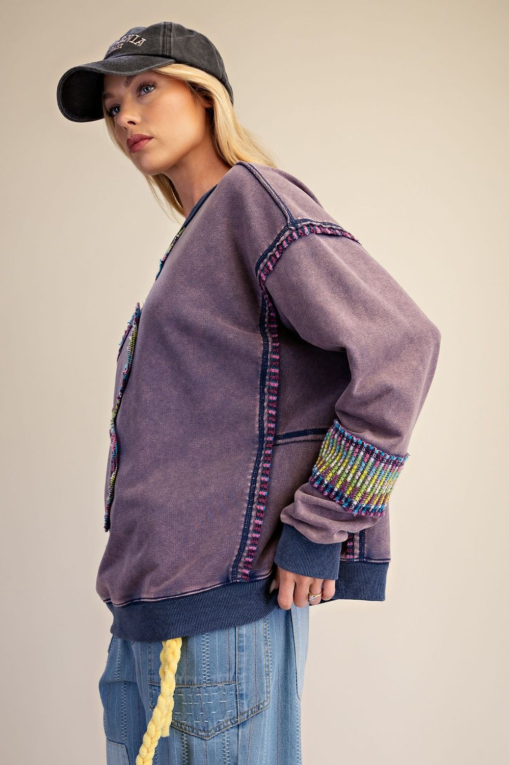 Side view of woman wearing dusty purple mineral wash oversized pullover showing colorful multicolor textured trim along seams and cuffs, with dropped shoulder silhouette