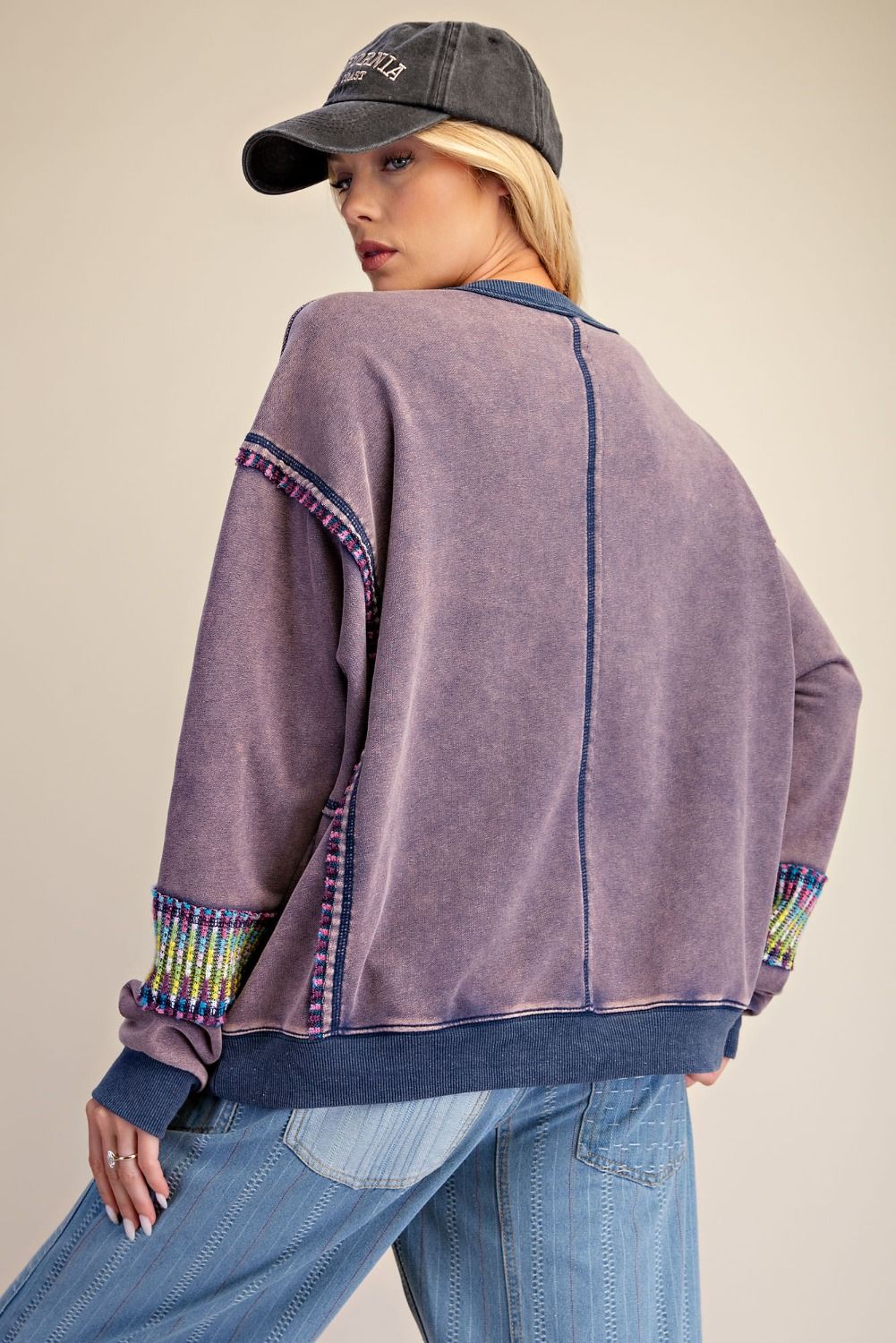 Back view of woman wearing dusty purple mineral wash crew-neck pullover with colorful trim cuffs and relaxed oversized silhouette, paired with wide-leg light wash jeans