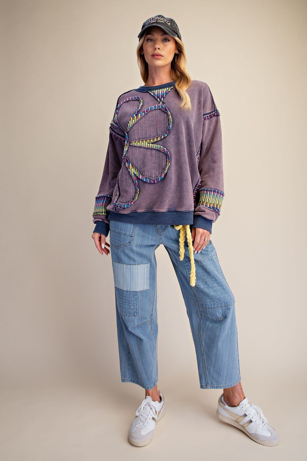Full-length front view of woman in dusty purple floral patch mineral wash pullover with colorful trim cuffs, styled with wide-leg light wash cargo pants, white sneakers, and a baseball cap