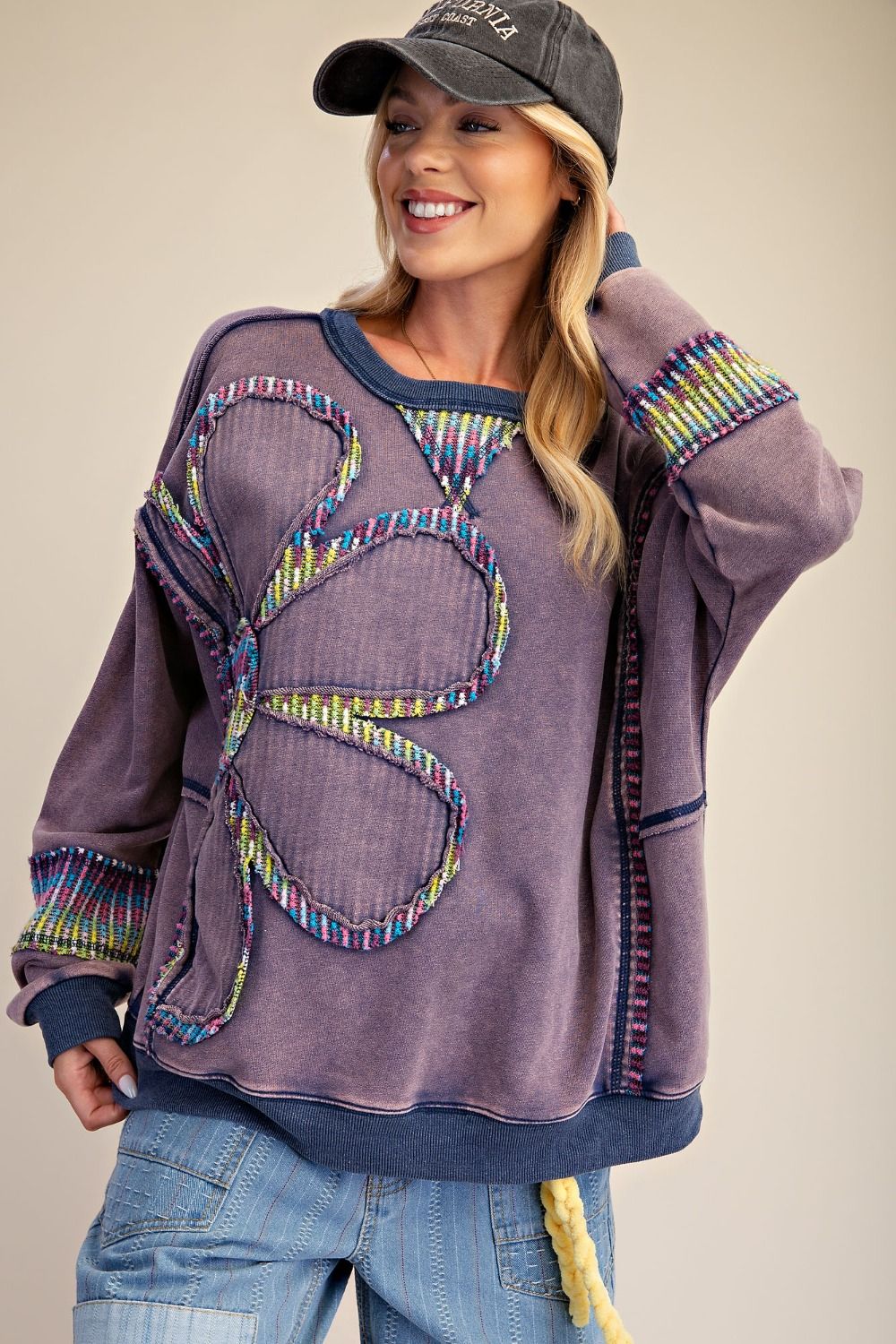 Close-up front view of woman wearing dusty purple mineral wash crew-neck pullover with large floral patchwork appliqué and colorful multicolor textured trim, paired with a baseball cap and wide-leg jeans