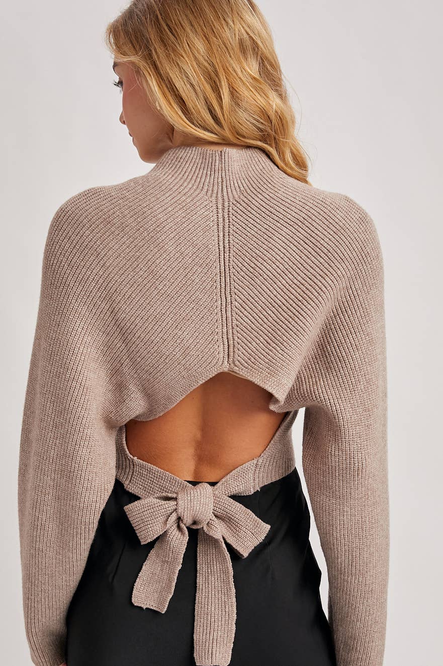 Back view of taupe ribbed open-back sweater with long sleeves and a waist tie bow, styled with a black skirt.