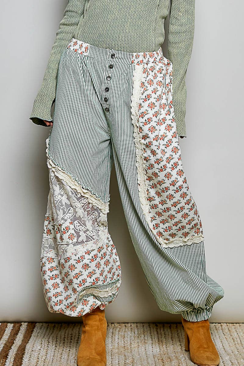 A person wearing loose, high-waisted patchwork pants with vertical panels of green-and-white stripes, floral prints, and lace trim, paired with a sage green sweater and tan ankle boots against a plain light wall.