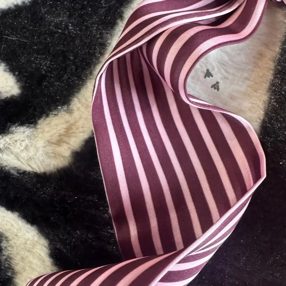 A close-up of a striped fabric accessory, likely a scarf or ribbon, featuring alternating vertical stripes of deep burgundy and light pink, laid out on a black and cream patterned faux fur surface.
