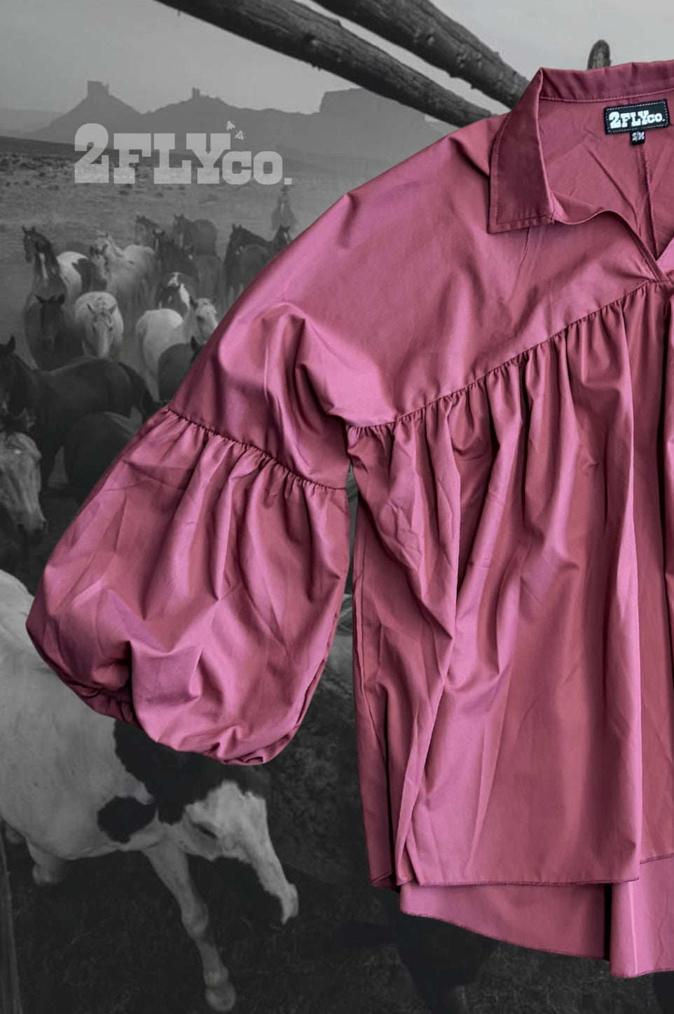 A dusty rose pink long-sleeve blouse with a ruffled yoke and voluminous puff sleeves, shown against a black and white background of a cowherd with a rustic wooden fence.