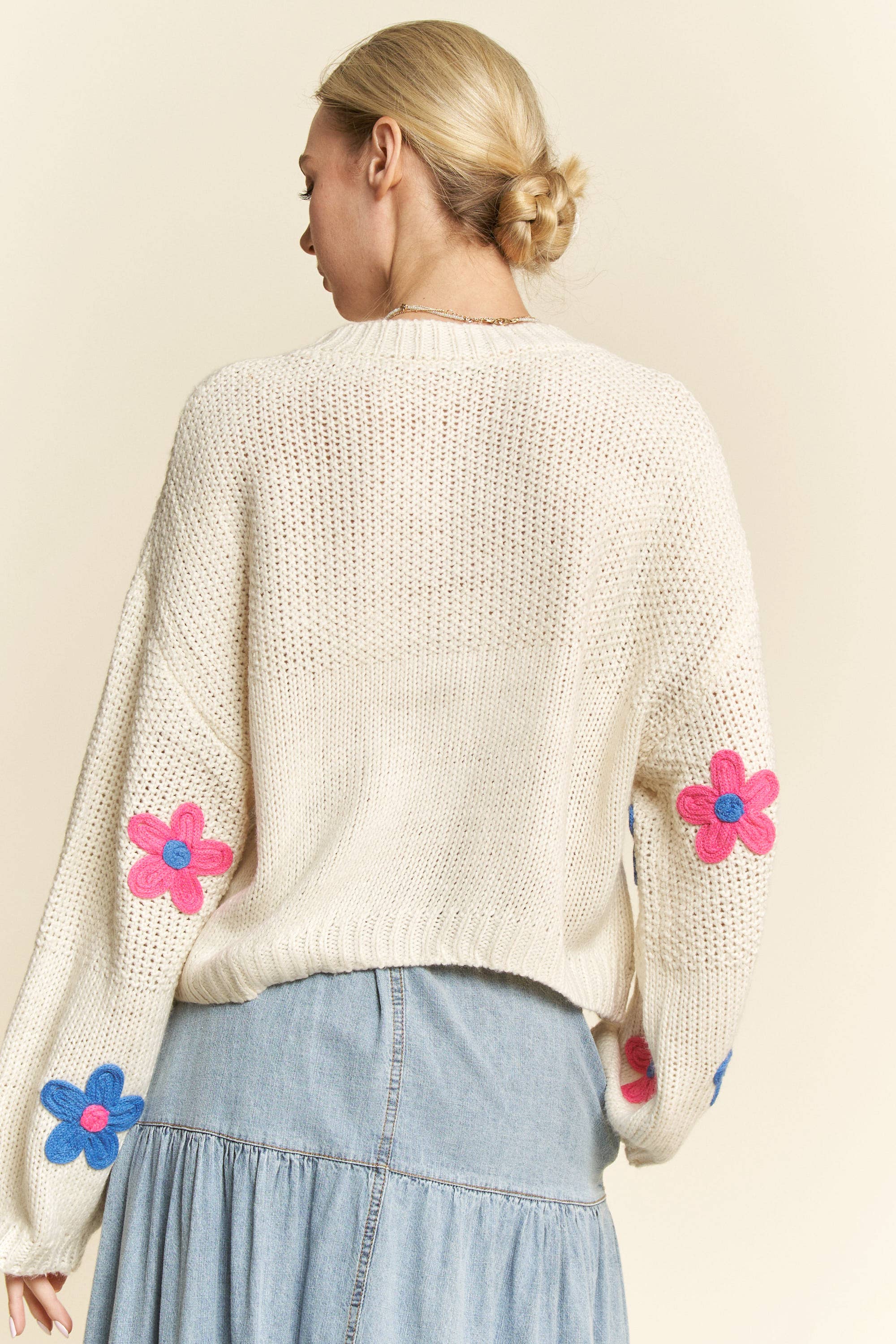 Back view- Cream knit sweater with colorful 3D floral appliqués in pink, blue, and green, styled with a button-front light denim skirt.