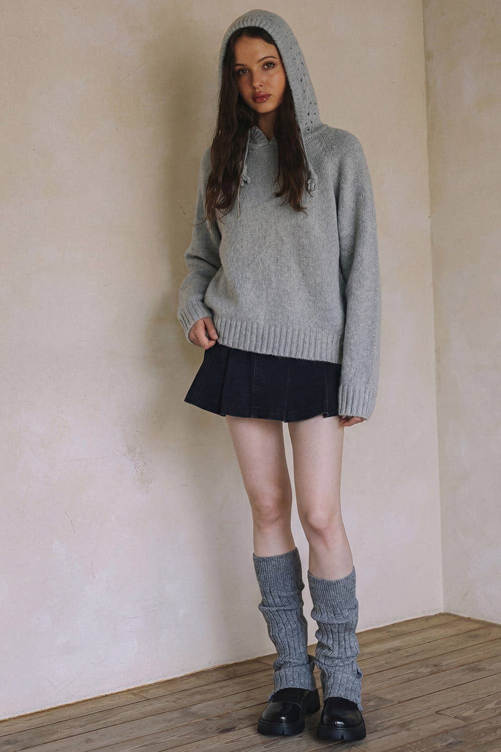 Woman wearing a gray sweater with a hood, black skirt, and gray knee-high socks against a plain wall.