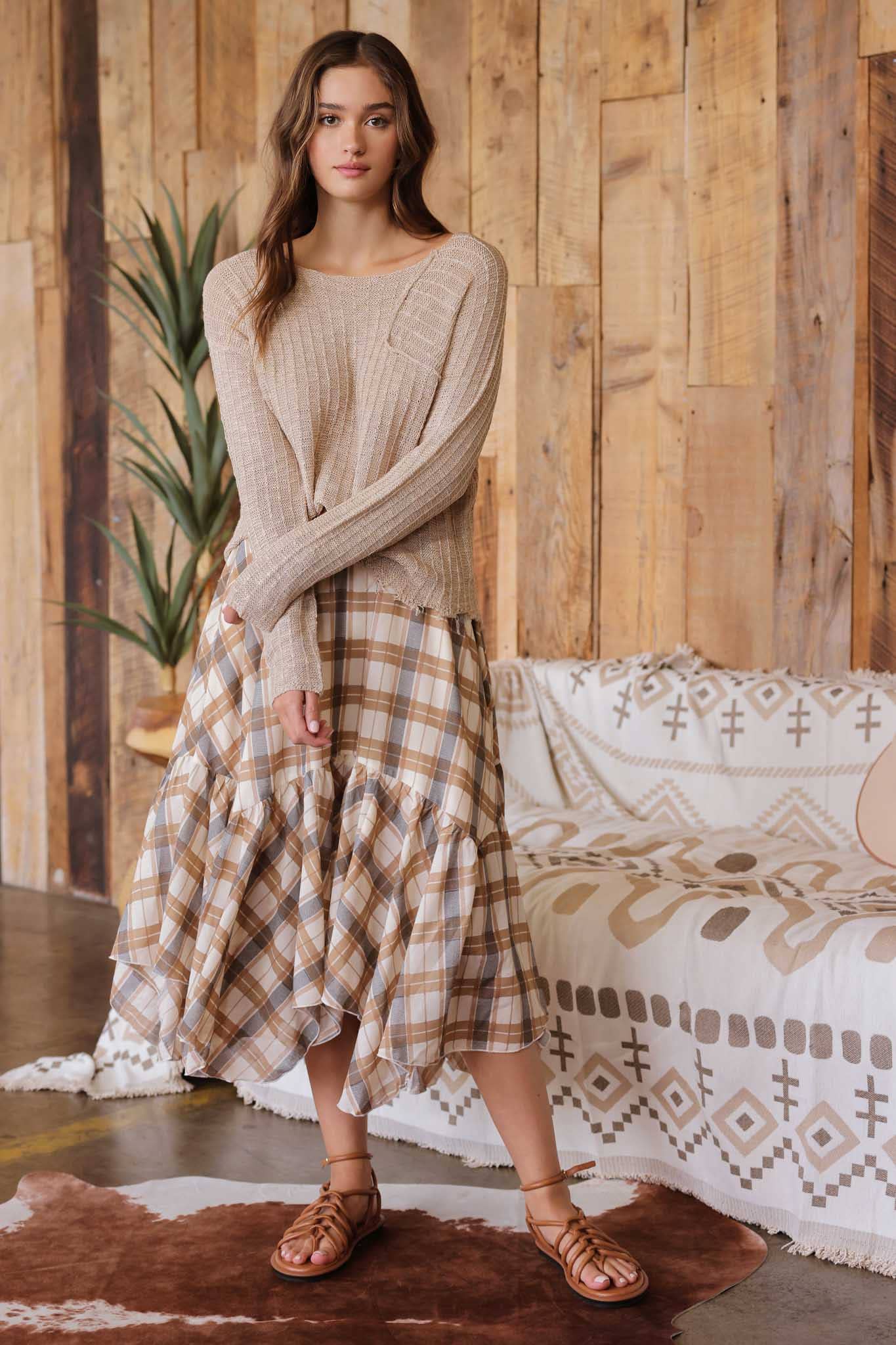 Front view  of model wearing taupe distressed lightweight sweater with ribbed knit texture, raw edge hem, and relaxed fit paired with tan plaid ruffle skirt