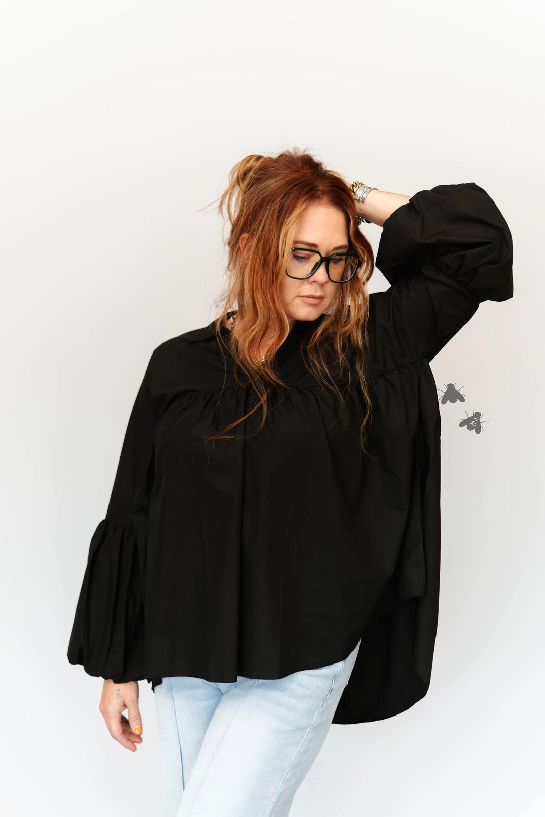 A woman with red hair and glasses wearing a black long-sleeve blouse with dramatic puff sleeves and light blue jeans, posing against a plain white background.