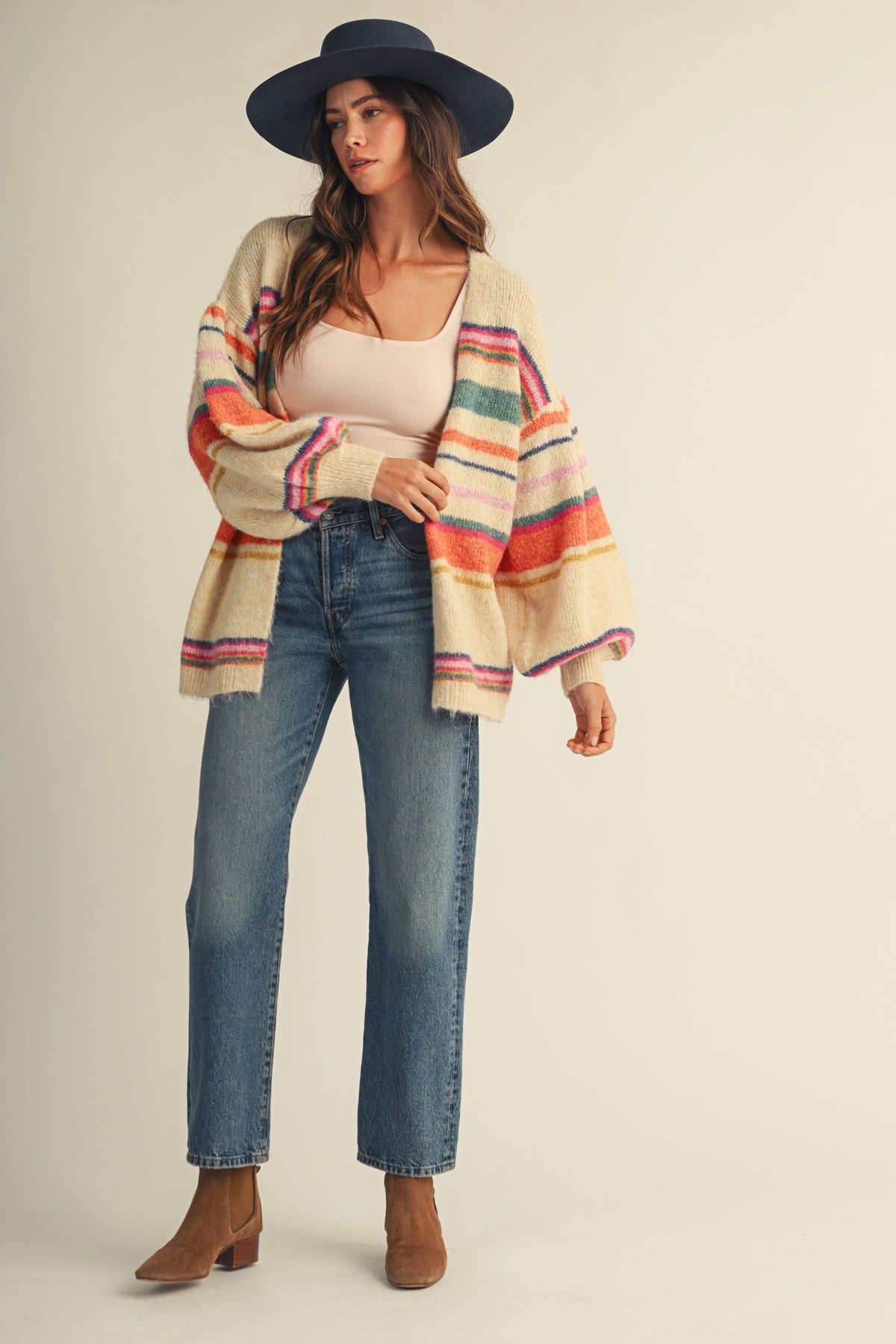 A woman modeling a cream knit cardigan with colorful horizontal stripes in orange, green, pink, and blue, paired with a black wide-brim hat, beige tank top, blue jeans, and brown ankle boots against a solid beige background.