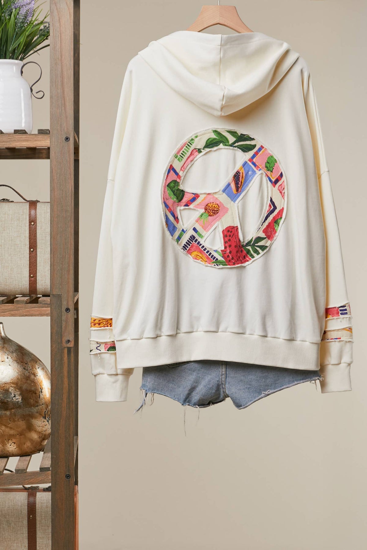An off-white hooded sweatshirt hanging on a wooden hanger, shown from the back with a large colorful circular graphic featuring a peace sign and tropical leaves. The hoodie is styled over light-wash denim shorts, set against a neutral beige wall with a rustic wooden shelf on the left.